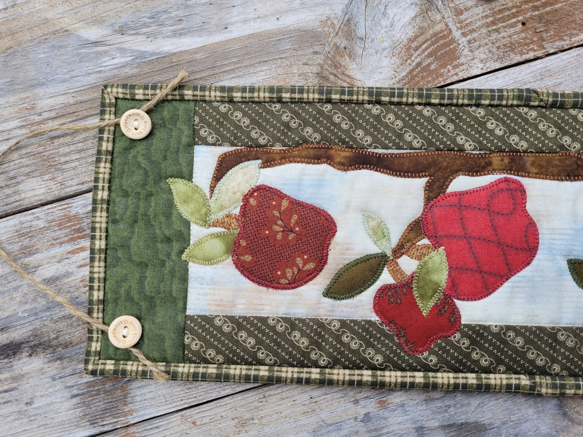 Skinny wall quilt is laid horizontal to show the top portion of the quilt with two wooden hanging buttons holding the jute string and the wonky red applique apples and tree branch on the quilt.