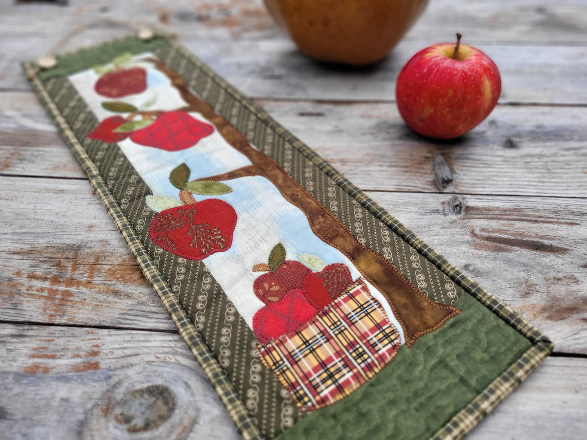 Angle view of a skinny wall quilt that features wonky red apples hanging from tree branches with a basket of apples underneath, shown on a weathered wood table with a red apple in the background.