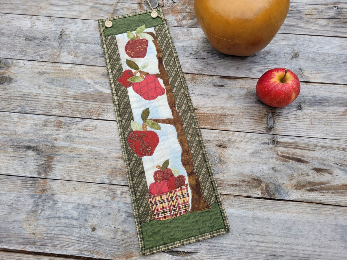 A narrow wall quilt featuring wonky red apples hanging from a tree branch with a plaid basket of apples underneath. Shown on a weathered wooden table with a red apple laying close by.
