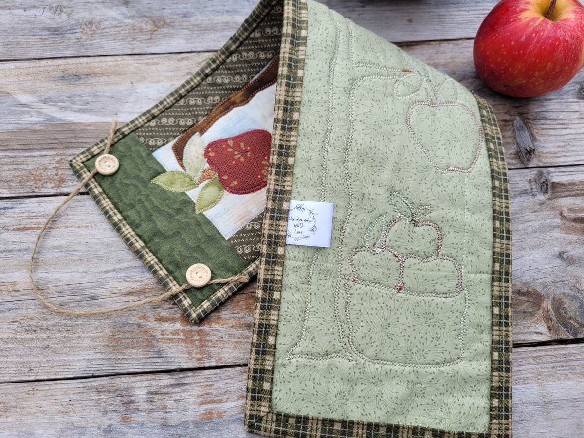 The narrow wall quilt is folded to show the green backing and stitches on a weathered wood table with red apple sitting nearby on the upper right.
