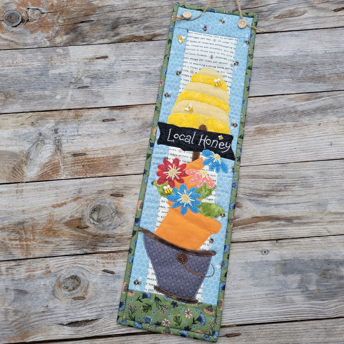 A long narrow quilt featuring a bee skep, a sign saying 'local honey' and a wonky bucket of flowers is shown laying askew on a weathered wood table.