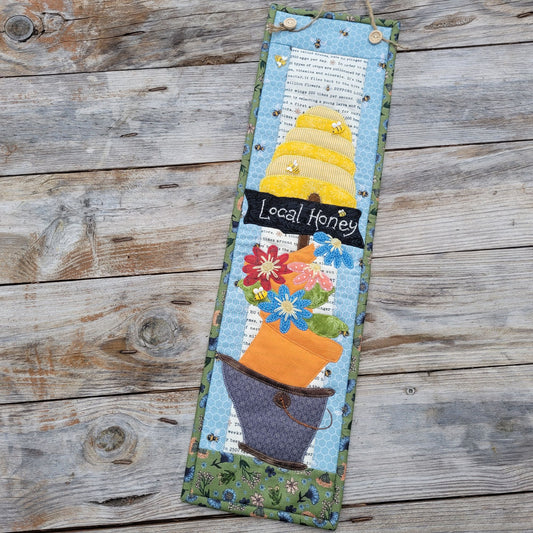 A long narrow quilt featuring a bee skep, a sign saying 'local honey' and a wonky bucket of flowers is shown laying askew on a weathered wood table.
