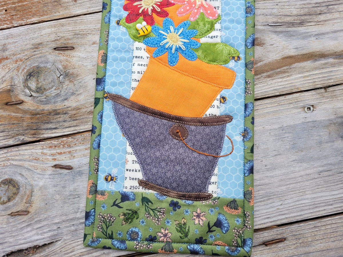 A close up of a narrow quilt with a wonky appliqued bucket holding a pot of daisy-like flowers, shown on a weathered wood surface.