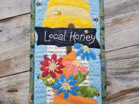 Close up of a narrow quilt showing a bee skep, an embroidered sign saying 'local honey' and some daisy-like flowers.
