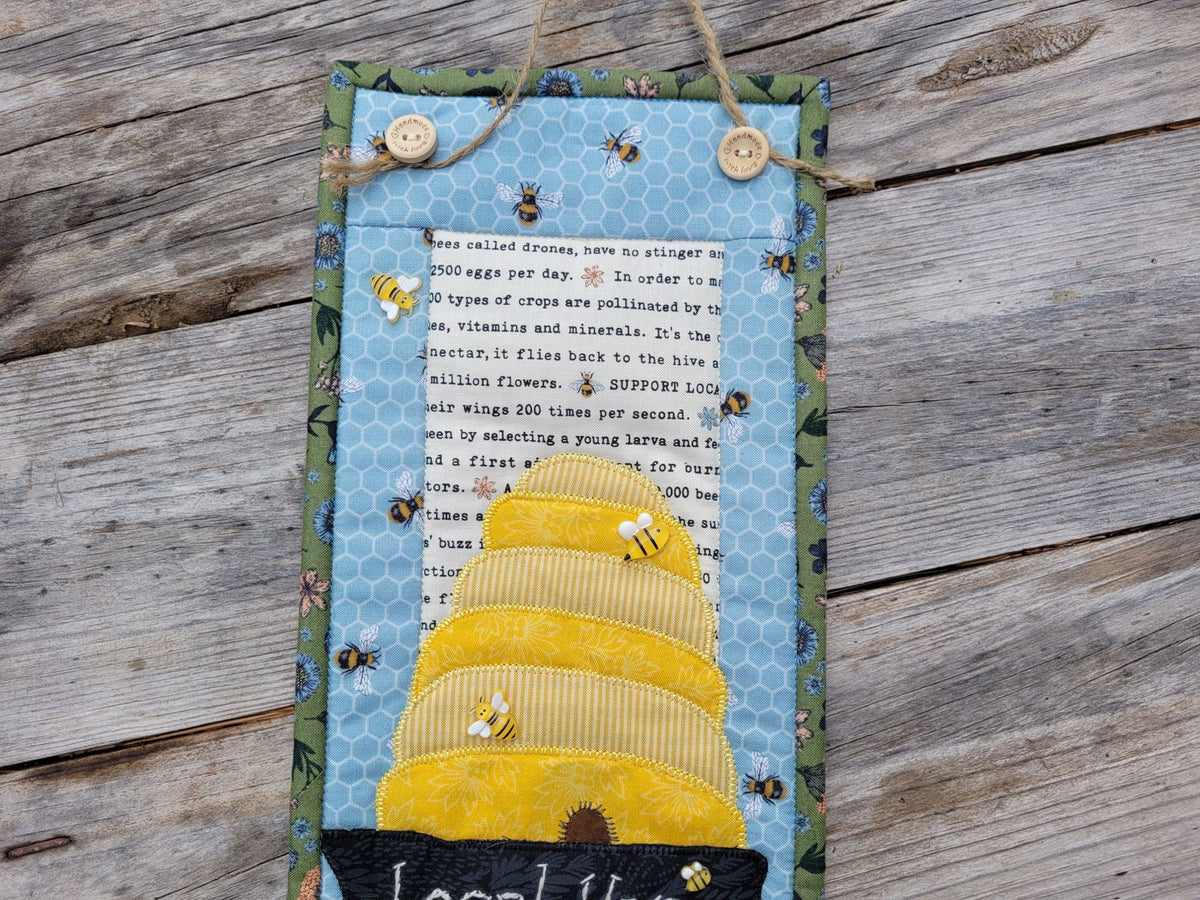 Close up of the top part of a narrow wall quilt featuring a bee skep with bee embelishments, and two wooden buttons holding the jute hanging string at the top. It is laid on a weathered wooden table.