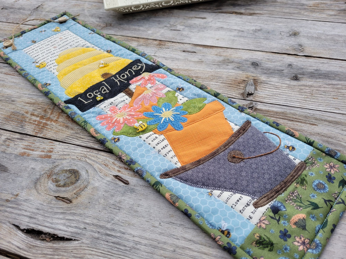 A skinny wall quilt featuring a bee skep, a sign saying 'local honey' and flowers in a bucket is laying diagonally across a weathered wooden table.