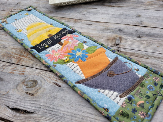 A skinny wall quilt featuring a bee skep, a sign saying 'local honey' and flowers in a bucket is laying diagonally across a weathered wooden table.