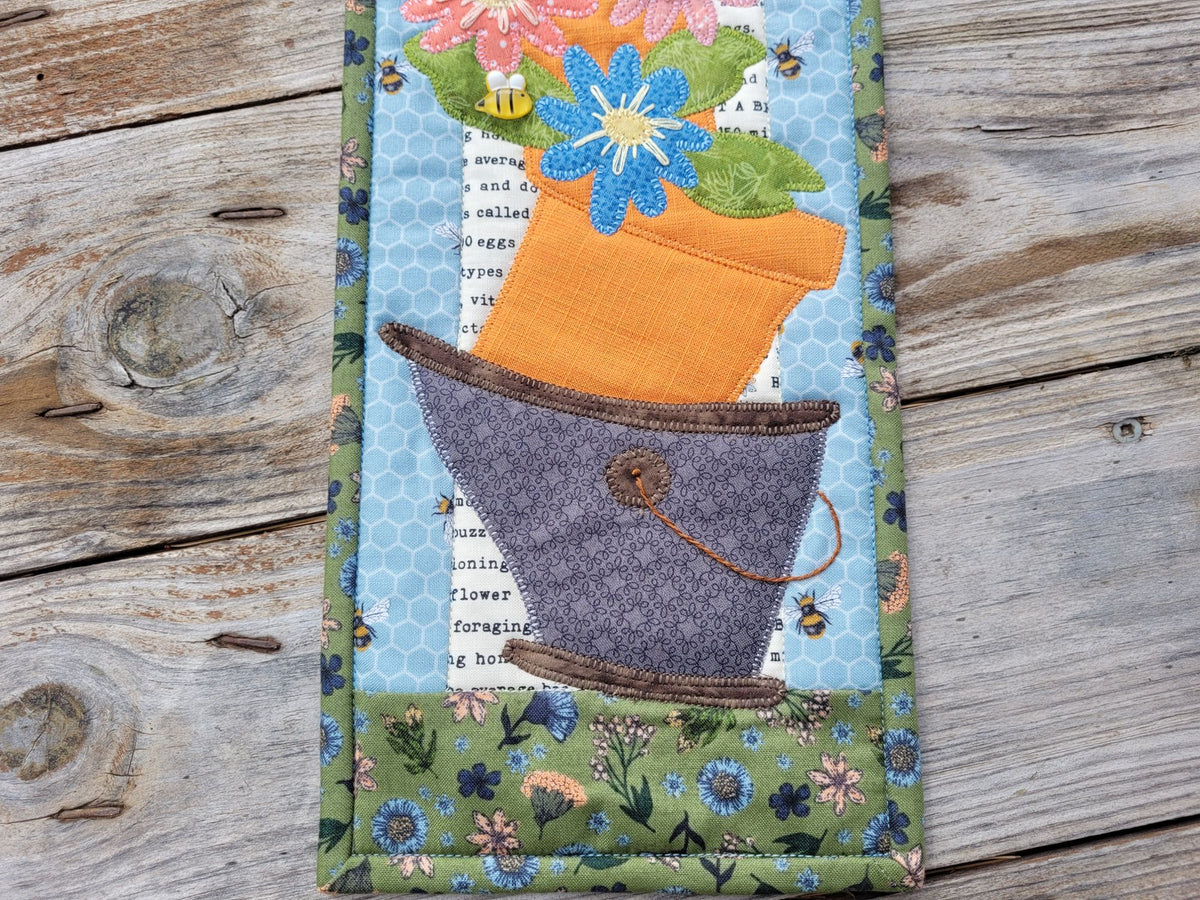Close up of the bottom half of the skinny wall quilt, showing the wonky bucket and pot of flowers, on a weathered wood surface.