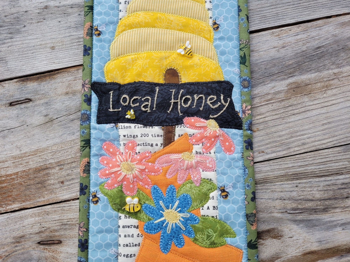 Close up of quilted wall hanging with a bee skep, a sign saying 'local honey' and several daisy like flowers, on a weathered wood table.
