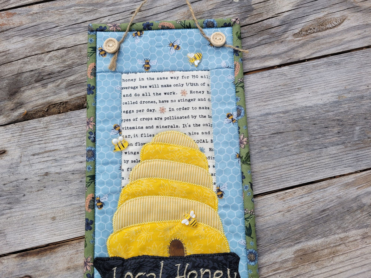 Close up of the top half of the skinny wall quilt, showing the bee skep with bee embellishments, on a weathered wood surface