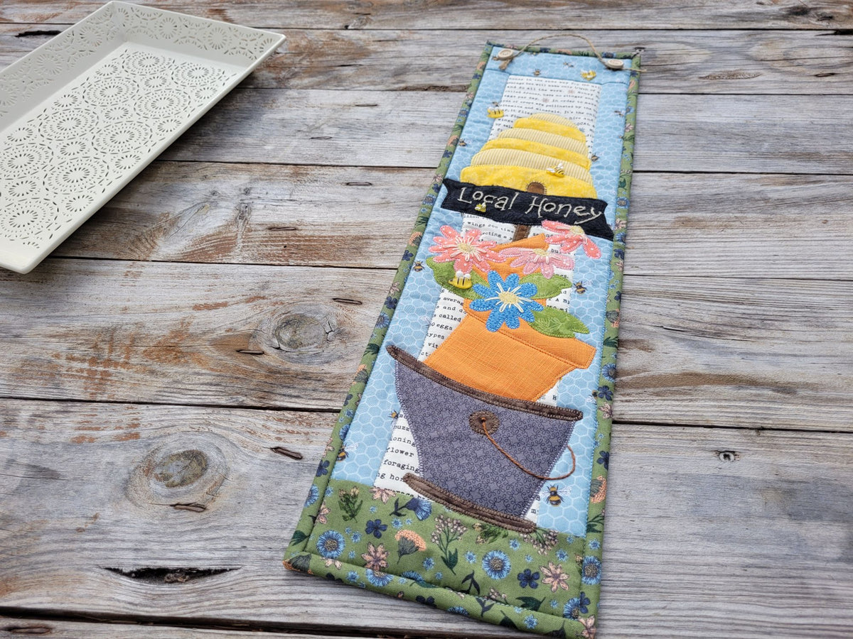 A narrow bee themed quilt is shown on a weathered wooden table with a white decorative tray partially visable on the left side.