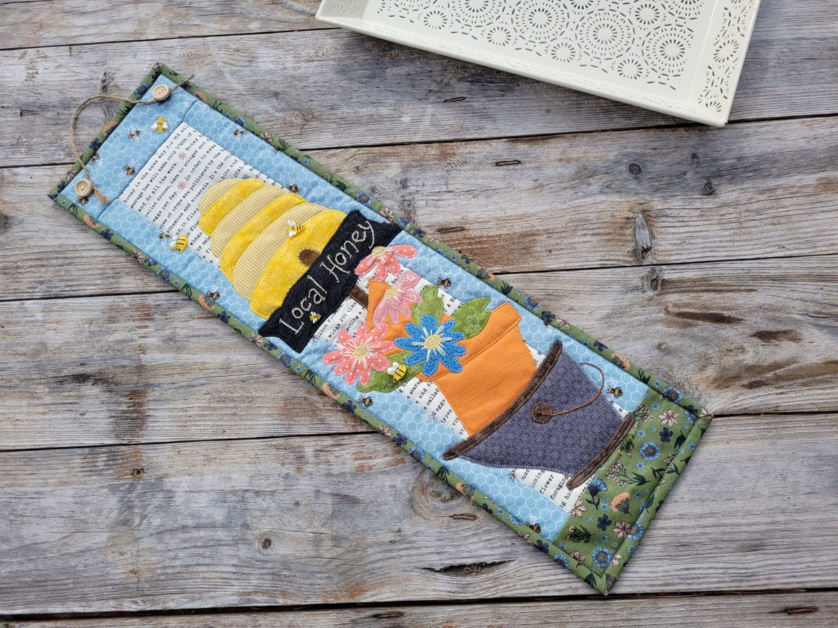 A skinny bee themed wall quilt is shown laying diagonally on a weathered wood table with a white decorative tray partially visable on the top.