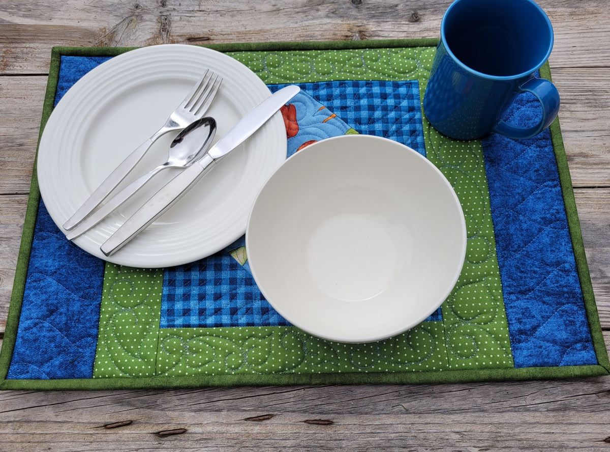 A quilted placemat with a green and blue gingham pattern, styled on a rustic wooden table with a white plate, silver cutlery, a white bowl, and a blue mug.
