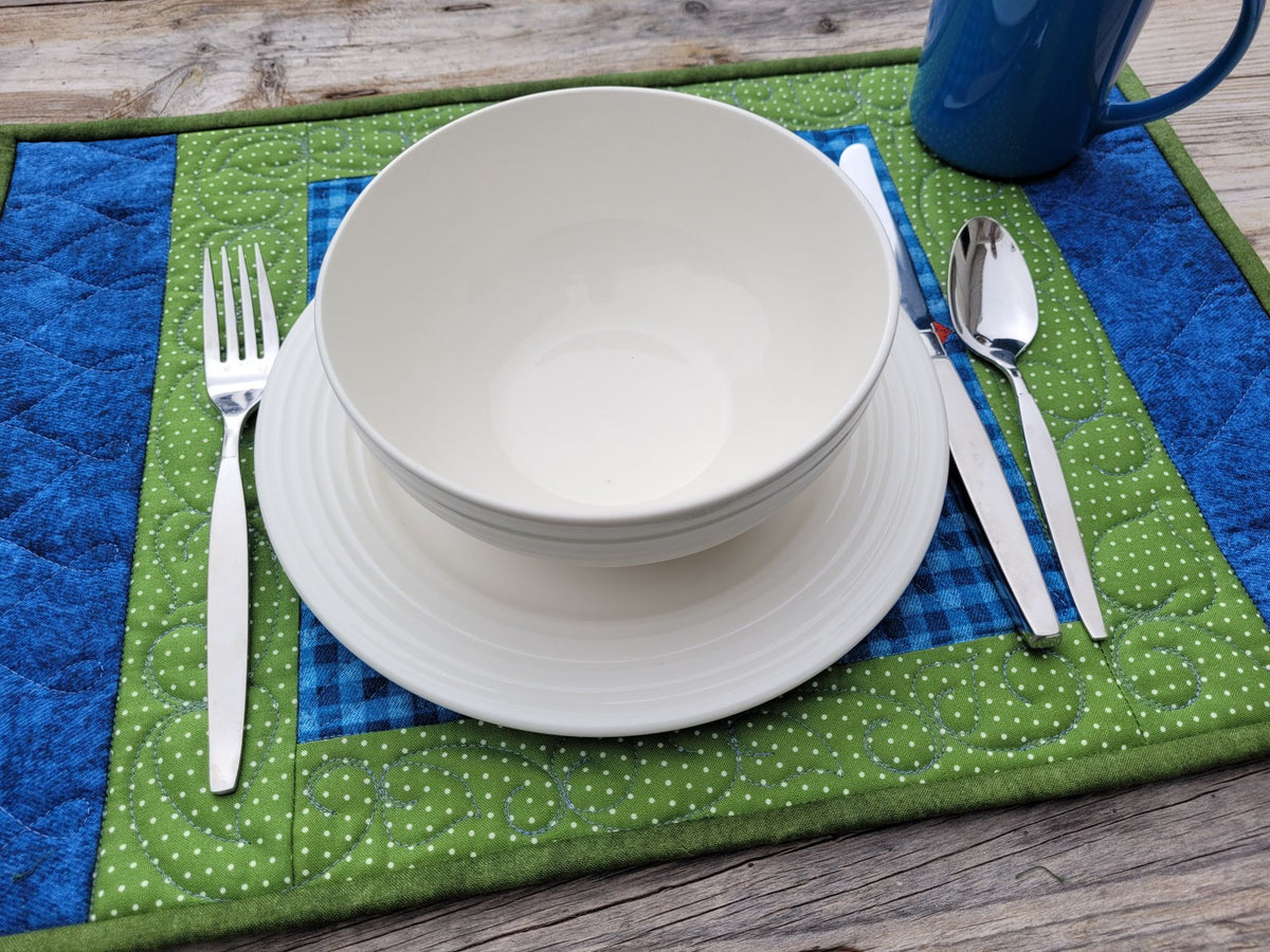 A place setting featuring a stack of white ceramic bowls and plates, silver flatware, and a blue mug, all arranged on a green and blue quilted placemat over a rustic wooden table.