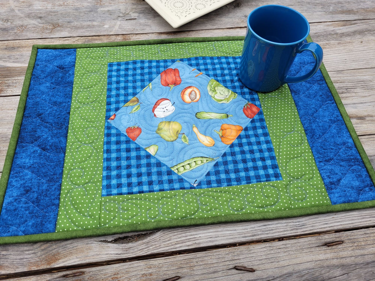 A handmade quilted placemat with concentric borders of green, blue gingham, and royal blue, featuring a central diamond patch with colorful fruit and vegetable illustrations. A blue ceramic mug sits on the placemat, which is laid out on a rustic wooden table.
