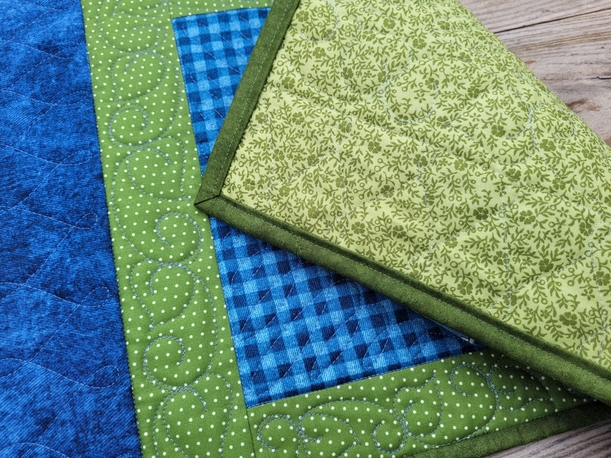 A close-up of a quilted placemat folded to show green floral backing. The quilt front is partially visable, showing some blue gingham center, and green dotted border, laid on a wood surface.