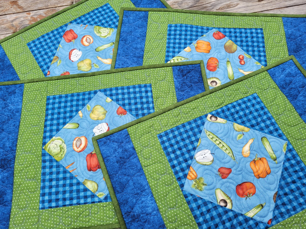 A set of quilted placemats with a colorful vegetable print, arranged on a rustic wooden surface. The placemats feature a blue and green gingham patterned center, a light blue background with a vegetable print, and a green dotted border on a green background.