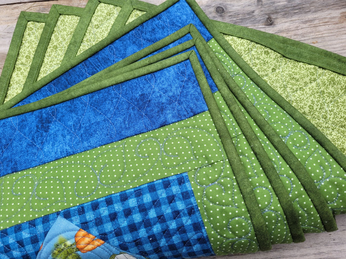 A set of handmade quilted fabric placemats in shades of green and blue, arranged in a fan-like stack on a rustic wooden surface.