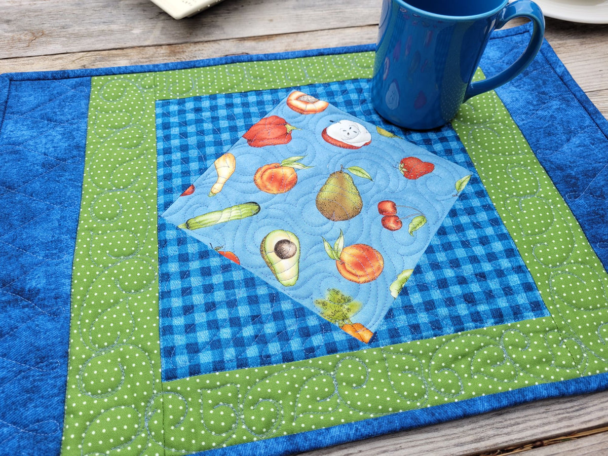A quilted fabric placemat with concentric borders in blue, green, and white polka dots, featuring a blue gingham section with a light blue fruit-and-vegetable center block and a blue mug resting in the corner, set on a wooden table.