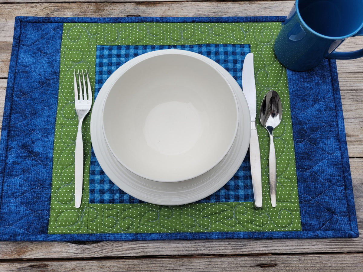 A place setting on a rustic wooden table featuring a blue and green quilted placemat, a stack of white ceramic bowls, silver cutlery, and a blue mug.