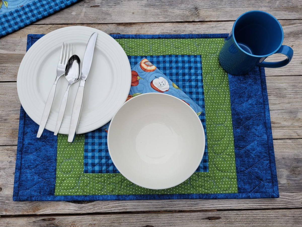 A styled tabletop place setting on a wooden surface featuring a quilted placemat in blue and green, a white plate with silver cutlery, a white bowl, and a blue mug.