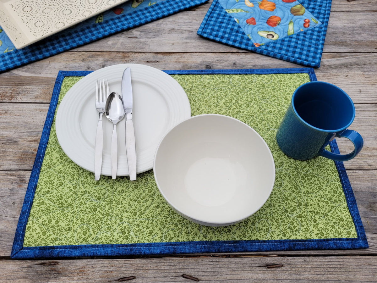 A green patterned placemat with a blue border is styled on a rustic wooden table with a white plate, silver cutlery, a white bowl, and a blue mug. Other quilts are partially visible in the background.