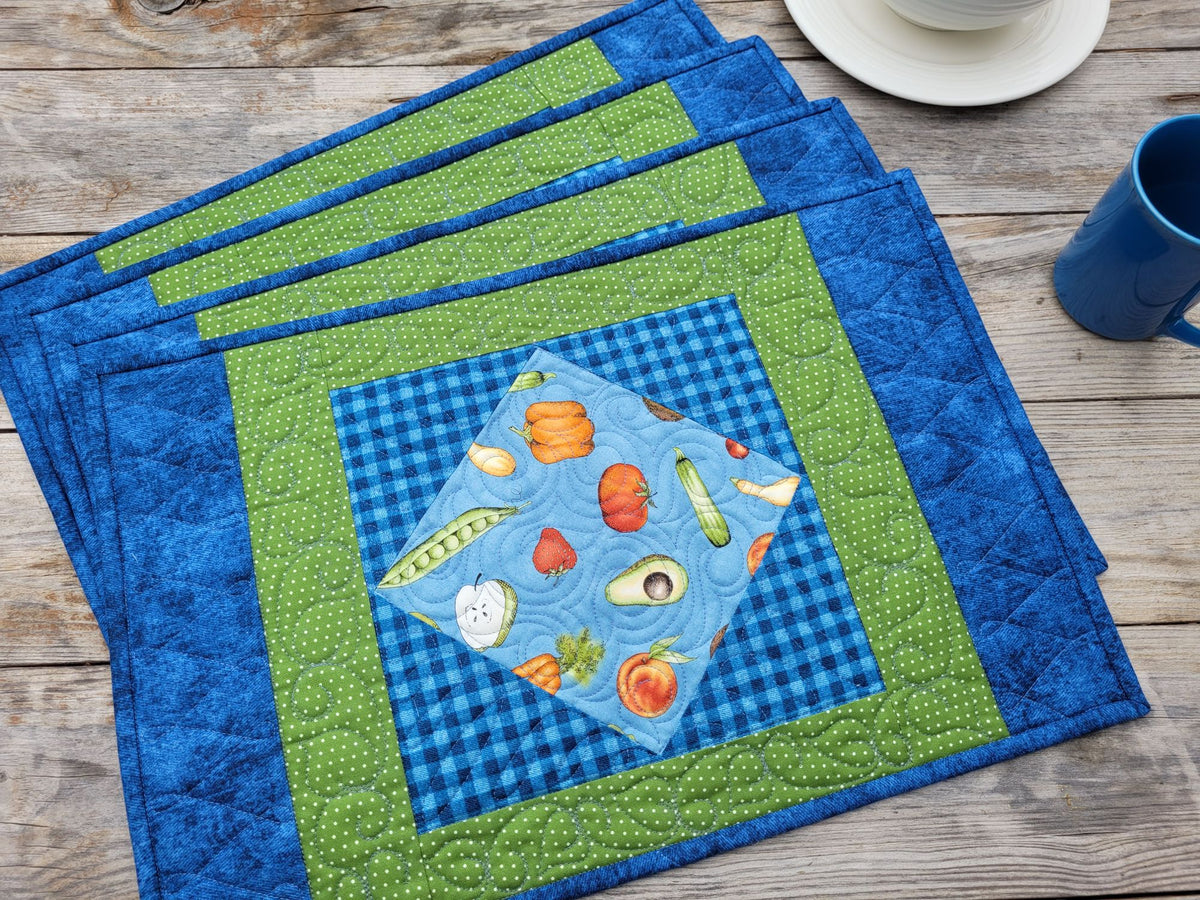 A set of four quilted fabric placemats arranged on a rustic wooden table. Each placemat features a central square with a vegetable-themed patchwork design on a light blue gingham background, surrounded by a green polka-dot border and a royal blue outer border.