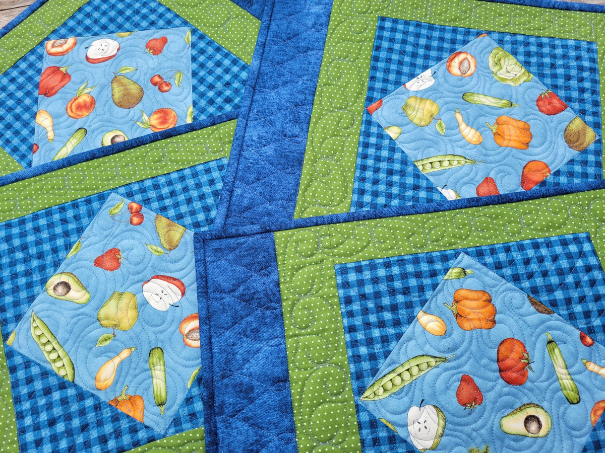 A close-up of a quilted placemat set of four. Each mat has a light blue background with a colorful vegetable print (peas, tomatoes, avocados, peppers, apples, etc) in the center, surrounded by a blue gingham border and a green dotted border.