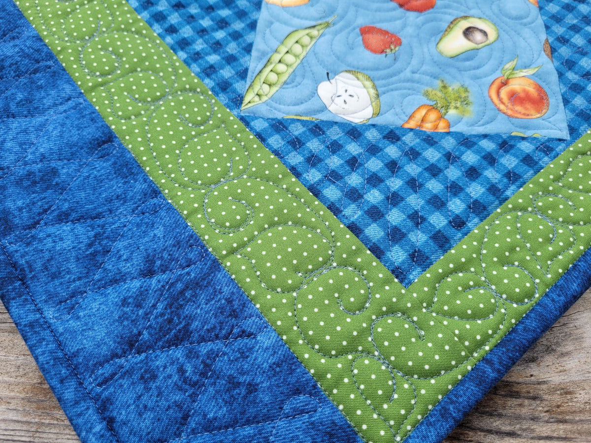 A close-up of a handmade quilted placemat, featuring a blue gingham check pattern, a green polka-dot border, and a central patch with a fruit and vegetable print on a light blue background.