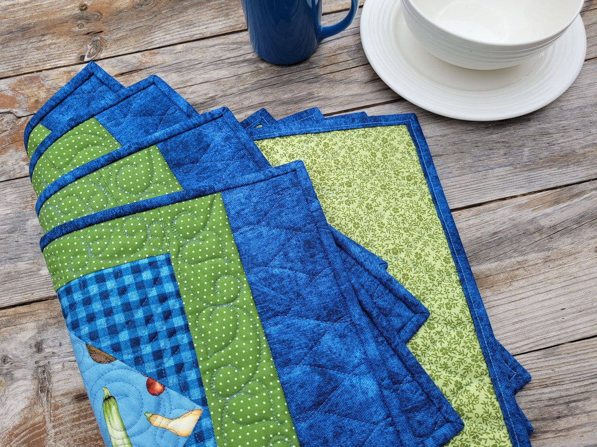 A set of handmade quilted placemats in shades of blue and green, featuring various patterns including gingham, polka dots, and floral prints, arranged on a rustic wooden table next to a blue mug and white bowl. Placemats are folded in a fanned out shape with the back partially visable.