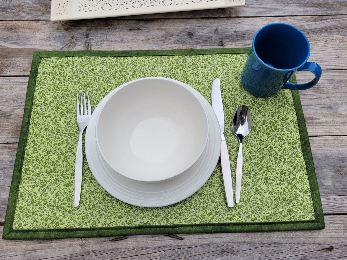 A place setting on a green patterned placemat with a white bowl, stacked plates, silver cutlery, and a blue mug on a rustic wooden table.