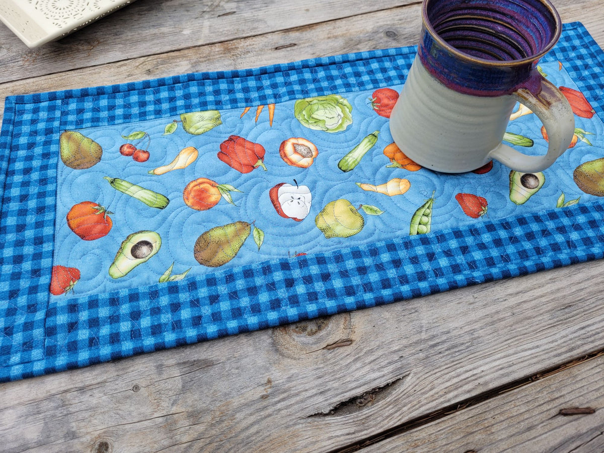 A quilted fabric table runner with a blue gingham border and a light blue center printed with colorful fruits and vegetables, shown on a rustic wooden table with a pottery mug resting on it.