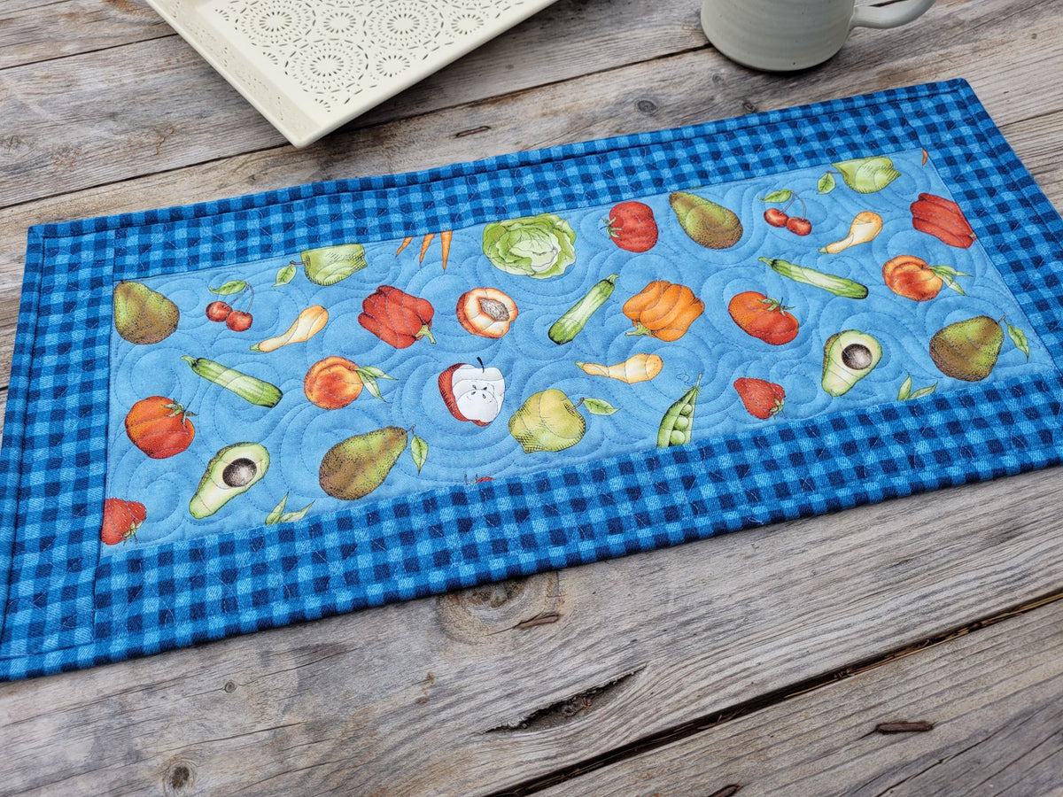 A rectangular quilted fabric table runner with a blue gingham border and a central light blue panel printed with colorful fruits and vegetables, laid on a rustic wooden tabletop.