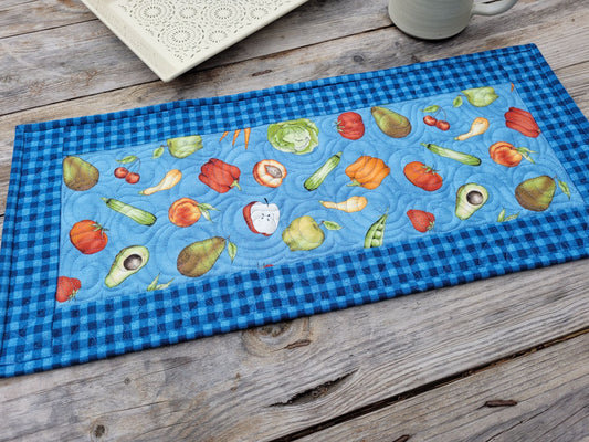 A rectangular quilted fabric table runner with a blue gingham border and a central light blue panel printed with colorful fruits and vegetables, laid on a rustic wooden tabletop.
