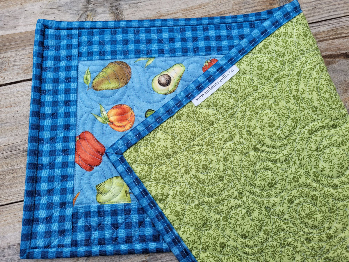 A quilted fabric table runner with a blue gingham border, a fruit print panel, folded to show a green floral reverse side, laid flat on a rustic wooden surface.