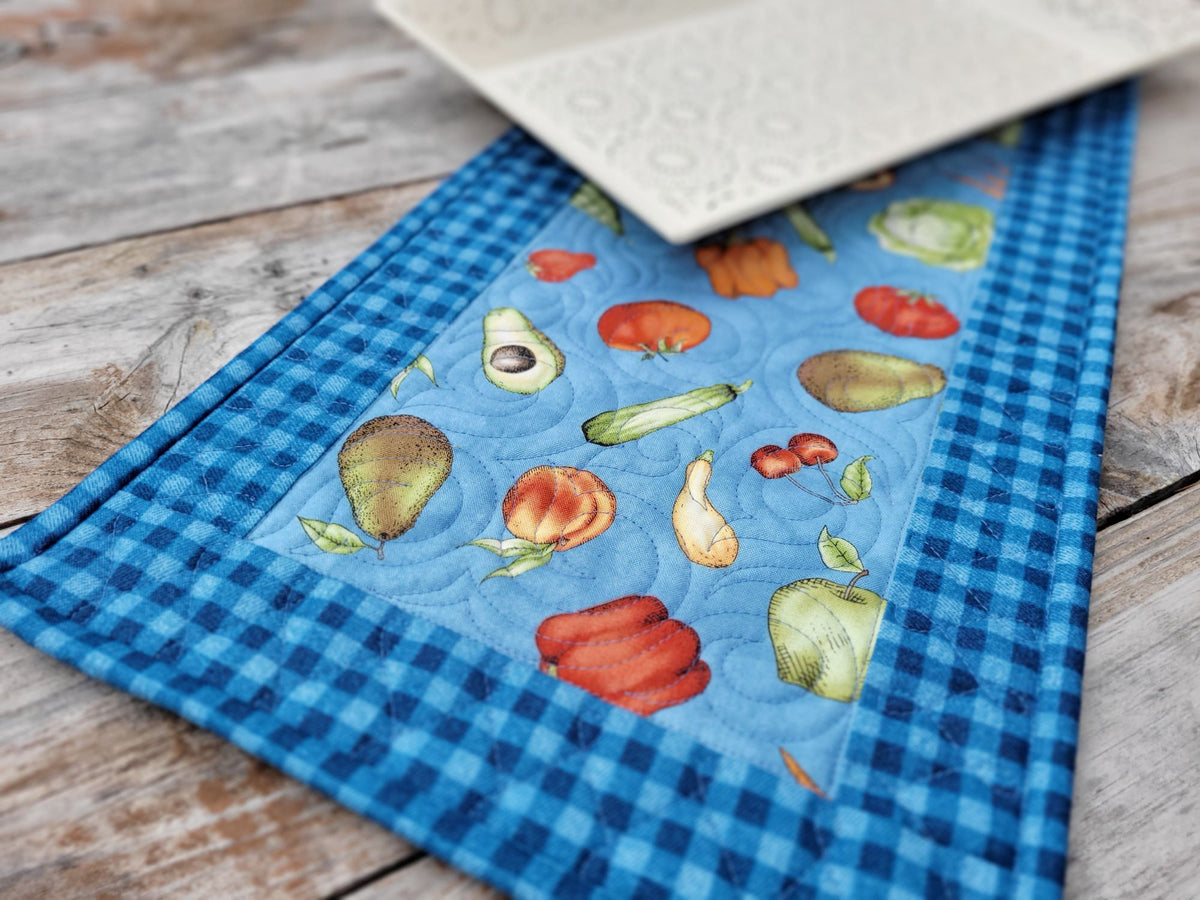 A square quilted fabric table runner with a blue gingham border and a central light blue panel featuring colorful fruit and vegetable illustrations, shown on a rustic wooden tabletop with a white decorative tray partially visible.