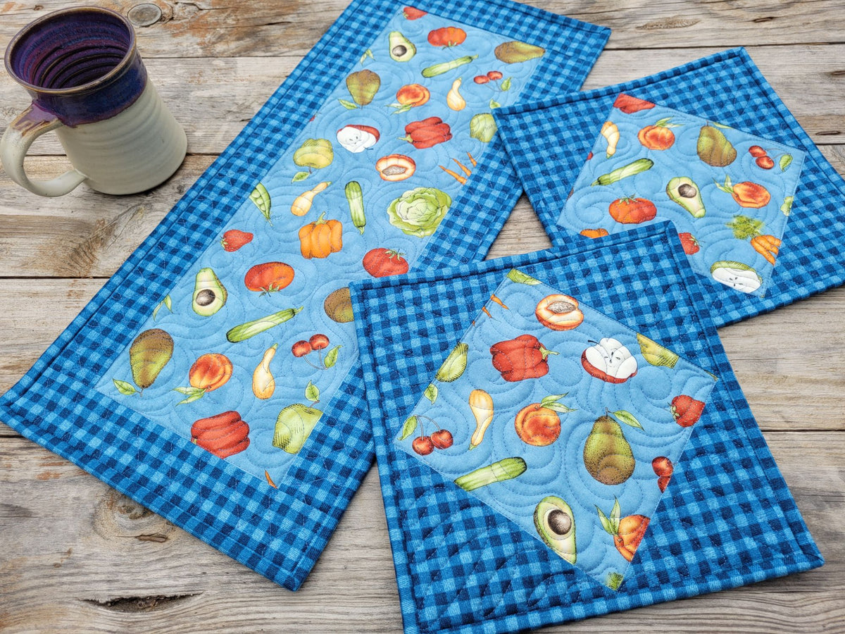 Three matching table quilts, one rectangle runner and two square hot mats, with blue gingham borders and a light blue center panel printed with colorful fruit and vegetable motifs, arranged on a rustic wooden tabletop next to a ceramic mug.