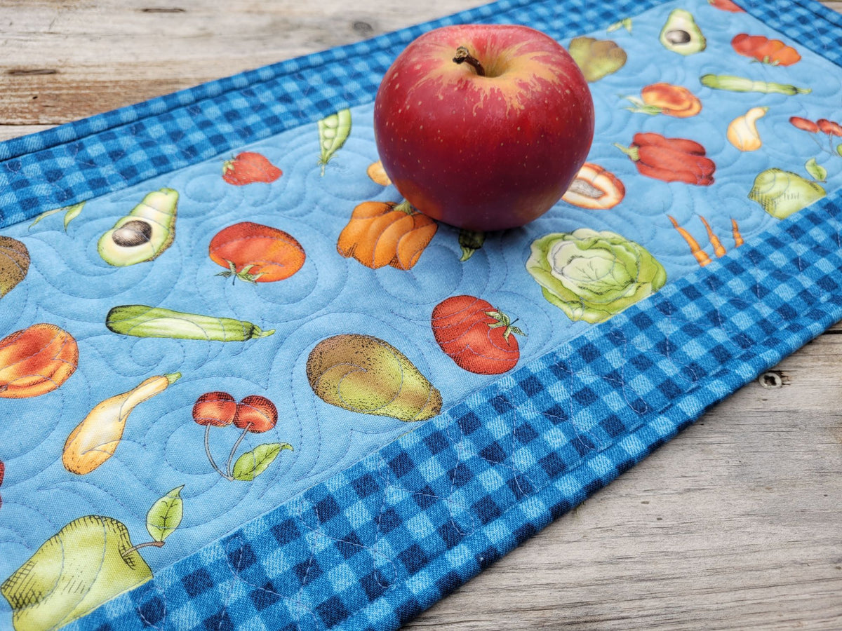 A quilted table runner with a blue gingham border and a light blue center featuring a colorful print of fruits and vegetables, shown on a rustic wooden table with a red apple placed on top.