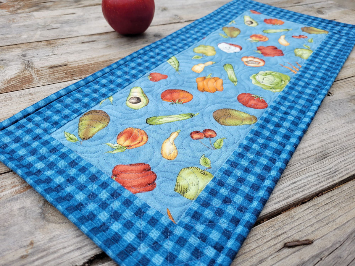 A rectangular quilted table runner with a blue gingham border and a central light blue panel printed with colorful vegetables, shown on a rustic wooden tabletop with an apple partially visible at the top.