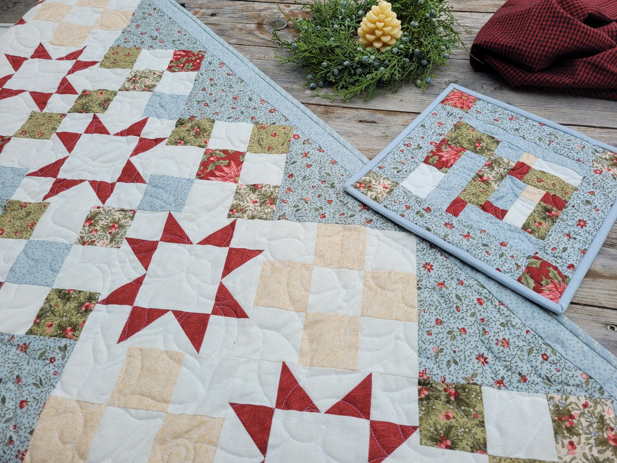 Christmas table runner and matching hot mat is shown on a weathered wooden table staged with greenery,  a candle and a red gingham towel.