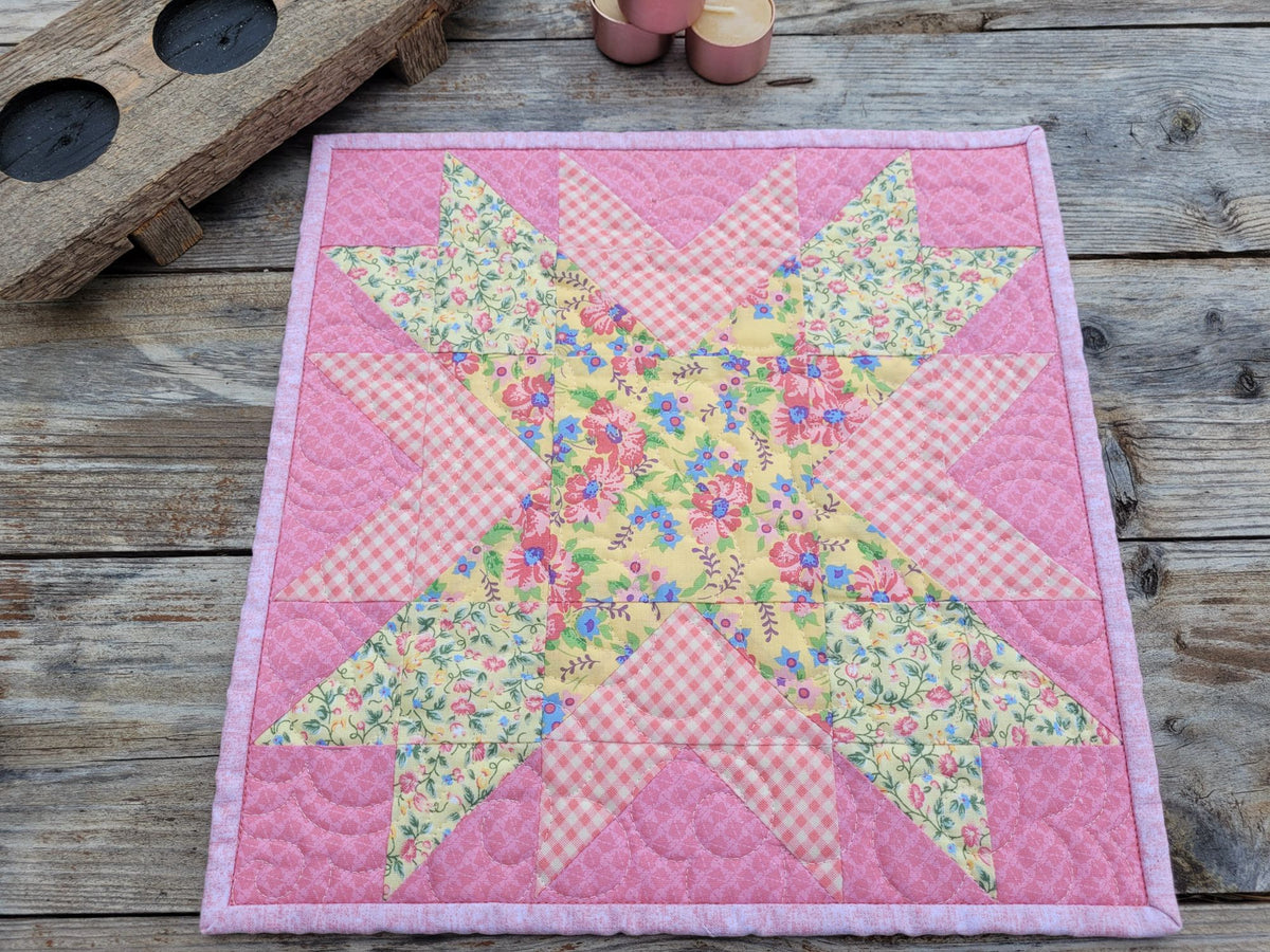 A small square patchwork quilt featuring a star pattern in yellow and pink floral fabrics sits on a weathered wooden table with candles partially visable in the upper left. 