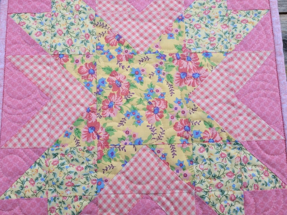 A small patchwork quilt featuring a star pattern done in yellow and pink floral fabrics with pink gingham fabric accent.