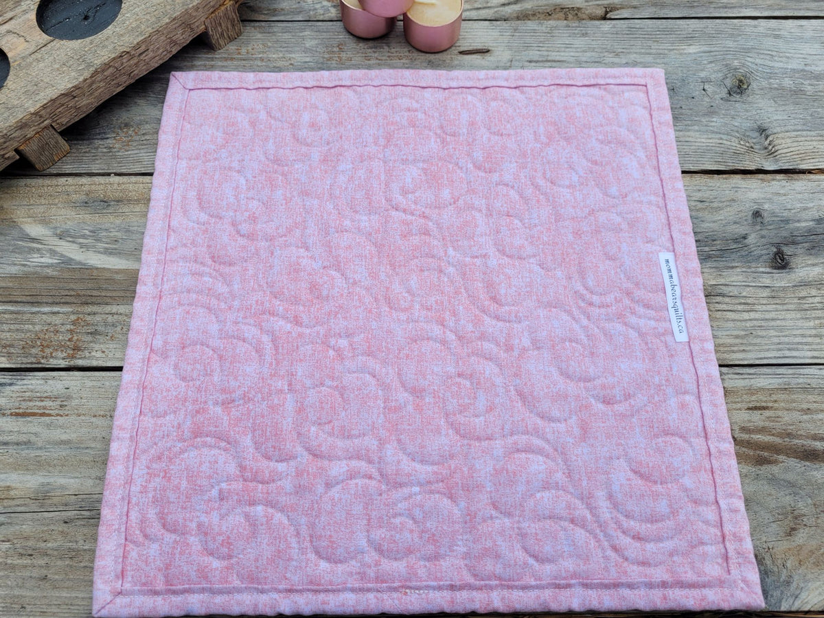 A small square quilt is laid on a weatherd wooden table with the back side showing a mottled pink fabric.