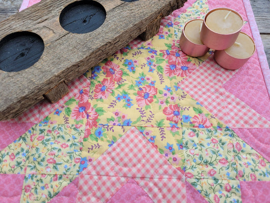 A wooden candle holder and a group of three beeswax tealight candles sit on a pink and yellow patchwork quilt with star shape pattern.