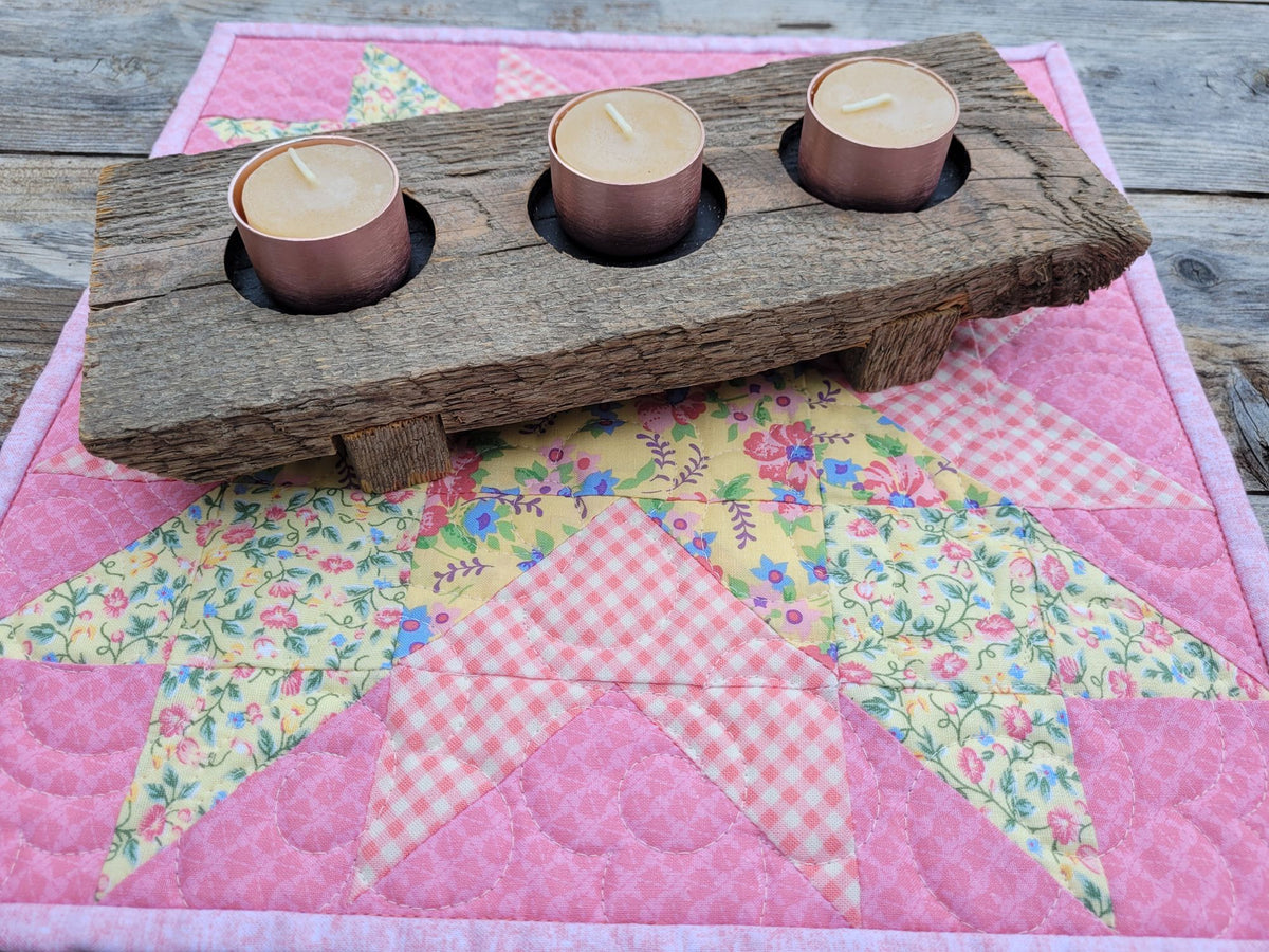 Quilted candle mat in pink and yellow floral  patchwork set on a weathered wooden table with a rustic wooden candle holder holding three beeswax tea light candles.