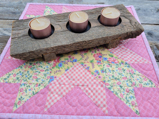 Quilted candle mat in pink and yellow floral  patchwork set on a weathered wooden table with a rustic wooden candle holder holding three beeswax tea light candles.