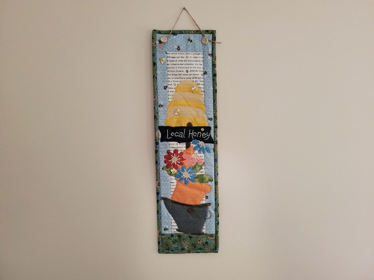 A skinny wall quilt featuring a bee skep, a sign saying 'local honey' and a bucket of daisy flowers is shown hanging on a plain beige wall.
