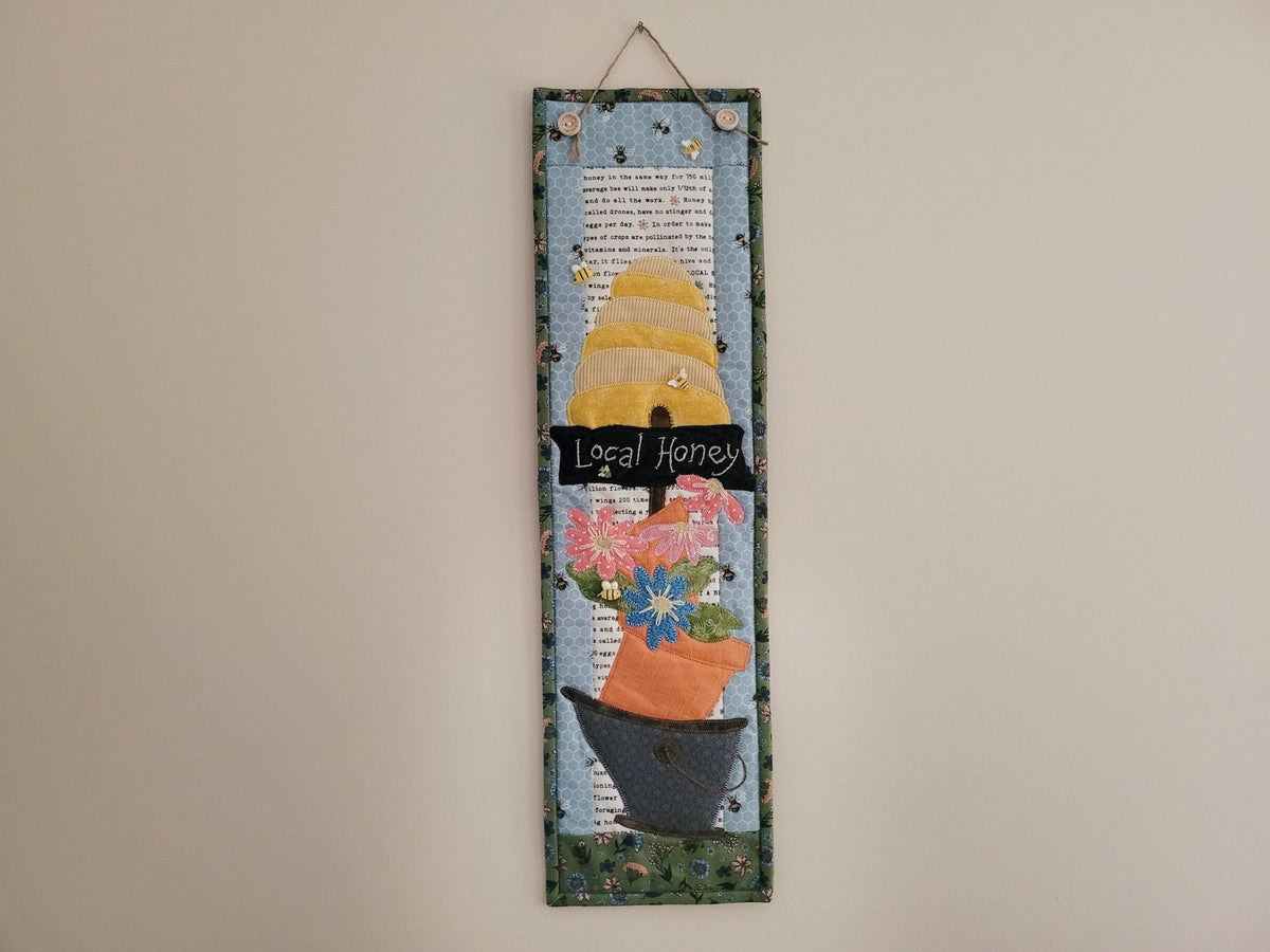 Narrow wall quilt featuring a bee skep, a sign saying 'local honey' and daisy-like flowers stacked in a wonky bucket is shown hanging on a plain beige wall.