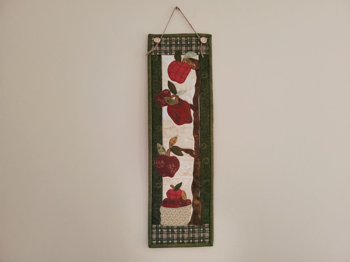A skinny apple themed wall quilt is shown hanging on a plain beige wall.