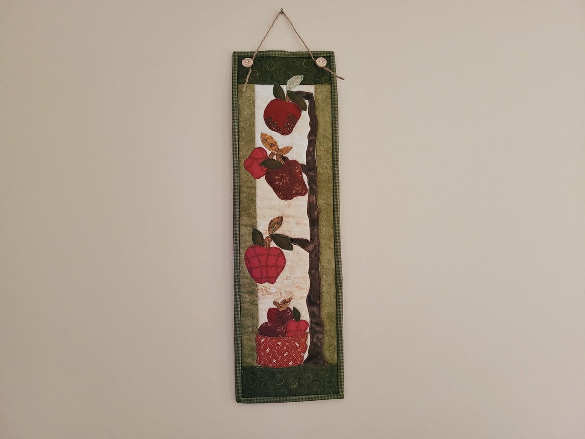 A skinny apple themed wall quilt is hanging on a plain beige wall.
